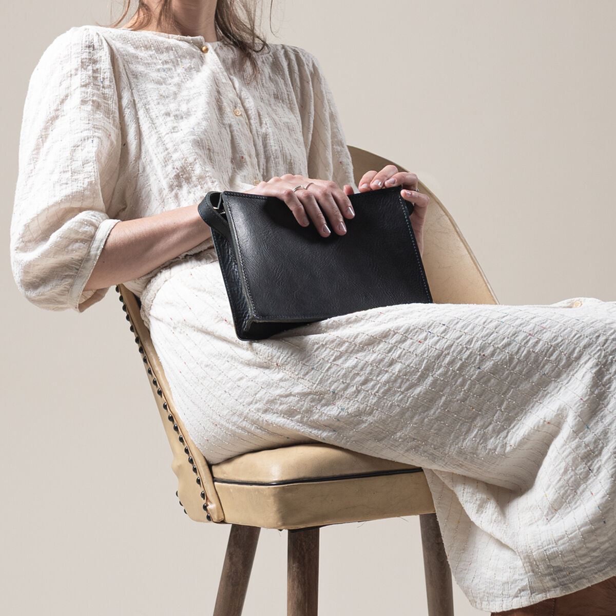 Eine Frau sitzt auf einem beigen Stuhl und hält eine schwarze Kernledertasche »Sas« auf ihrem Schoß. Sie trägt ein langes, cremefarbenes Kleid. Die Szene wirkt minimalistisch und elegant. Das Bild hebt die hochwertige Ledertasche als stilvolles Accessoire hervor.