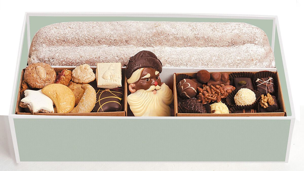Eine Geschenkbox mit verschiedenen Weihnachtsleckereien, darunter Lebkuchen, Pralinen, Dominosteine und ein Stollen, auf einem hellgrünen Hintergrund.