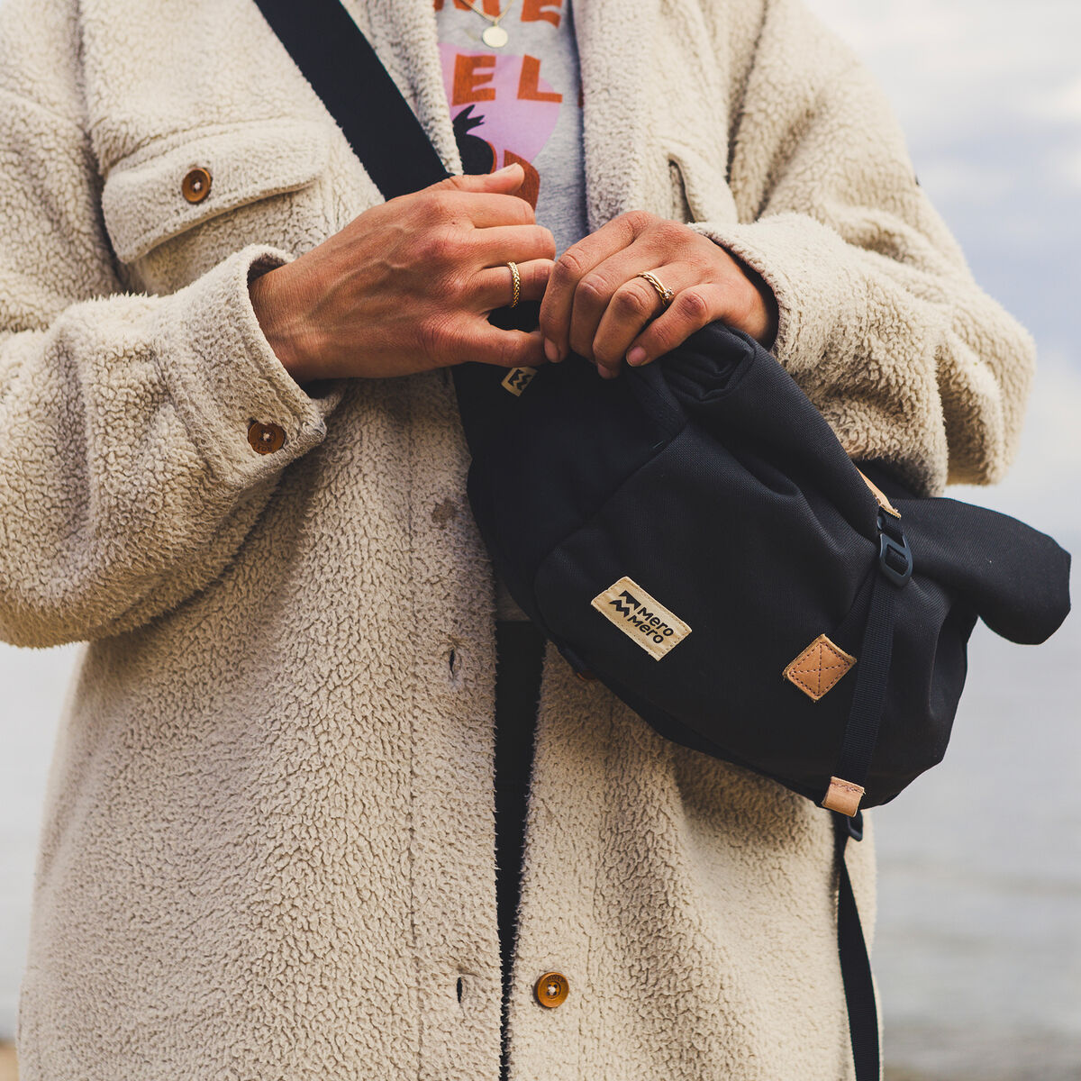 Eine Person trägt eine schlichte schwarze Umhängetasche »Piha« aus Segeltuch. Die Tasche wird über der Schulter getragen und hält durch robustes Design. Die Person trägt einen beigen Fleece-Mantel und einen weißen Pullover.