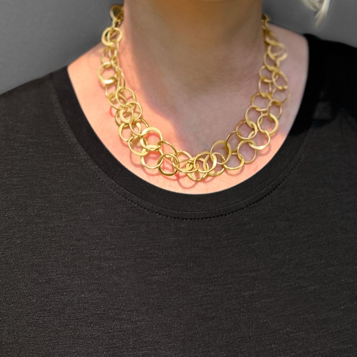 Eine Frau trägt ein »Double«-Collier aus vergoldetem Silber. Sie trägt eine schwarze Oberbekleidung und eine Brille und hat kurzes blondes Haar. Der Hintergrund ist grau.