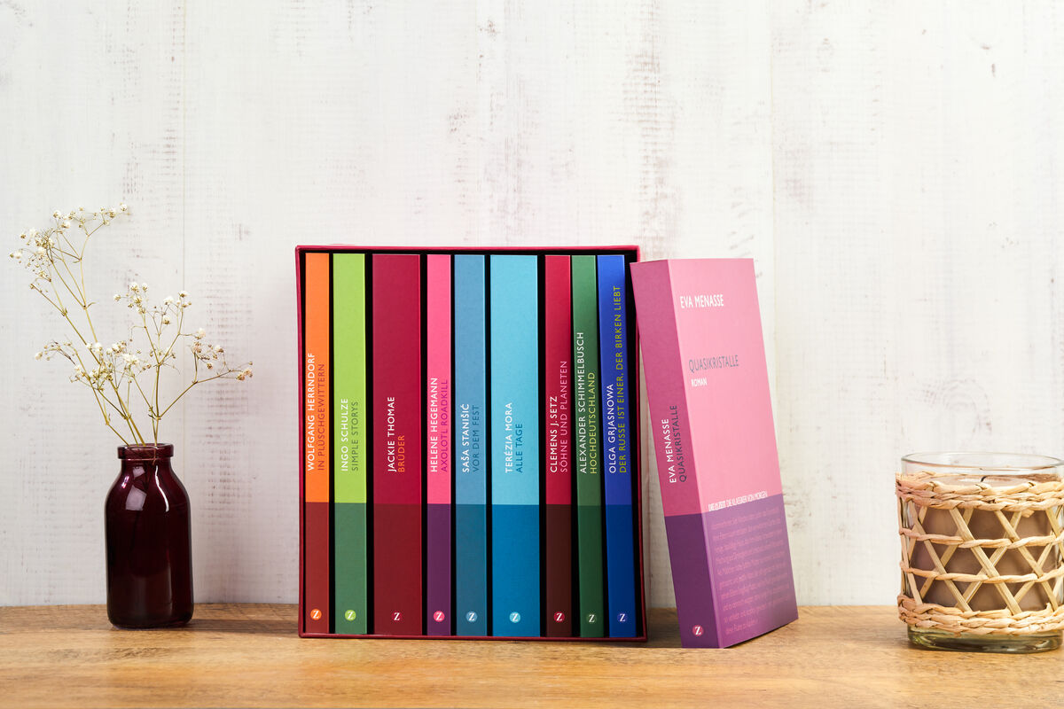Ein Set bunter Bücher der ZEIT-Edition »Die Klassiker von morgen« steht auf einem Holzregal. Daneben befindet sich eine kleine violette Vase mit dünnen weißen Blumen und ein Kerzenhalter mit einer beige geflochtenen Umhüllung.