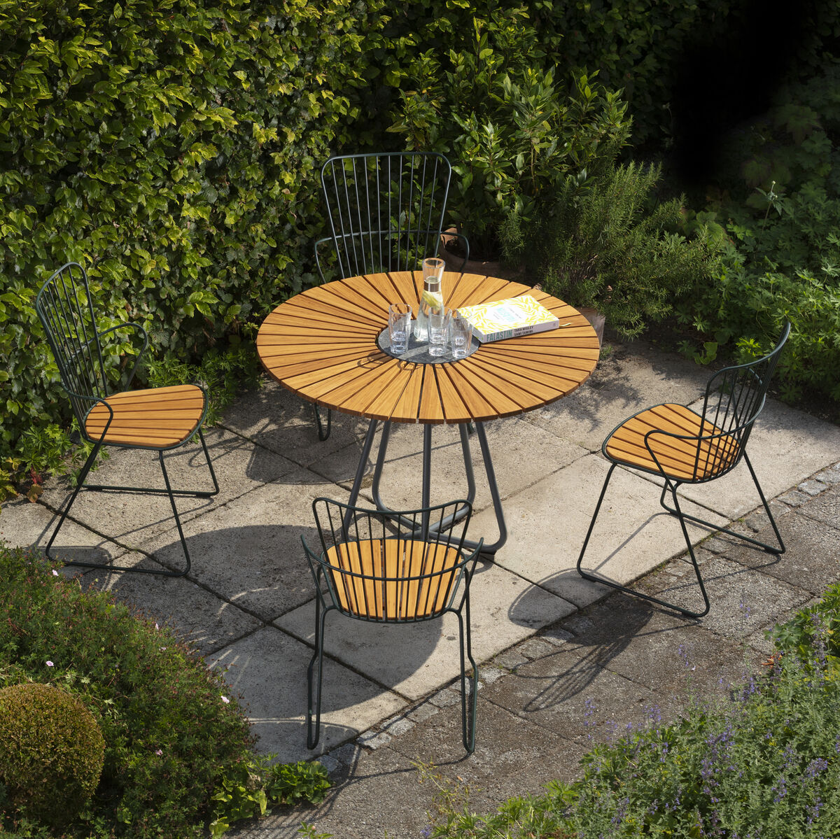 Ein stilvoller Essstuhl »PAON« steht draußen in einem Garten auf einem gepflasterten Platz. Der Stuhl hat eine schwarze Metallstruktur mit Holzlatten als Sitz- und Rückenlehne. Ein runder Tisch und andere ähnliche Stühle sind ebenfalls zu sehen, umgeben von grünen Büschen und Sträuchern.