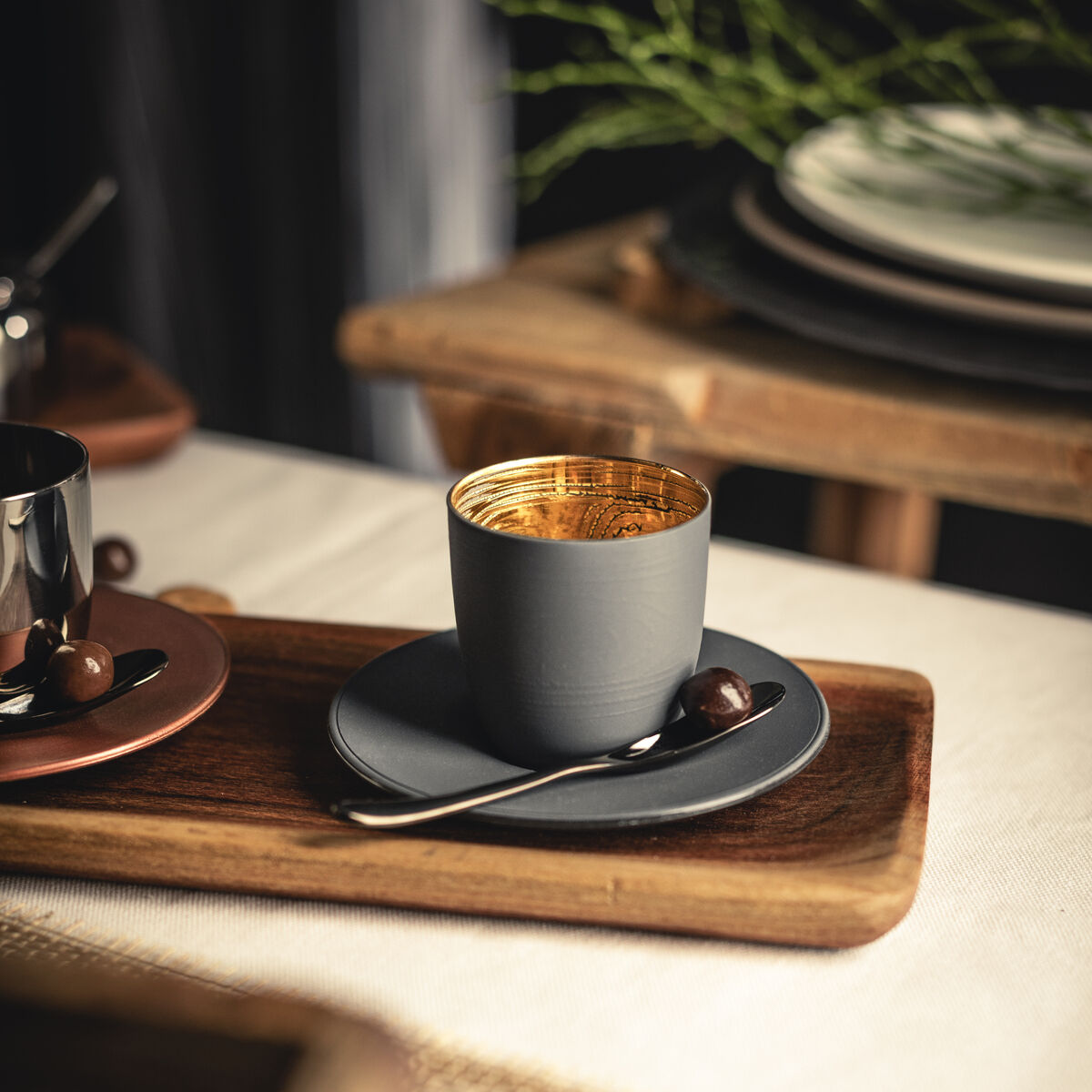 Espressoglas mit goldenem Innenrand und passendem Untersetzer steht auf einem rustikalen Holztablett, daneben ein silberner Löffel und eine Schokoladenpraline, arrangiert auf einem festlich gedeckten Tisch mit natürlicher Dekoration.