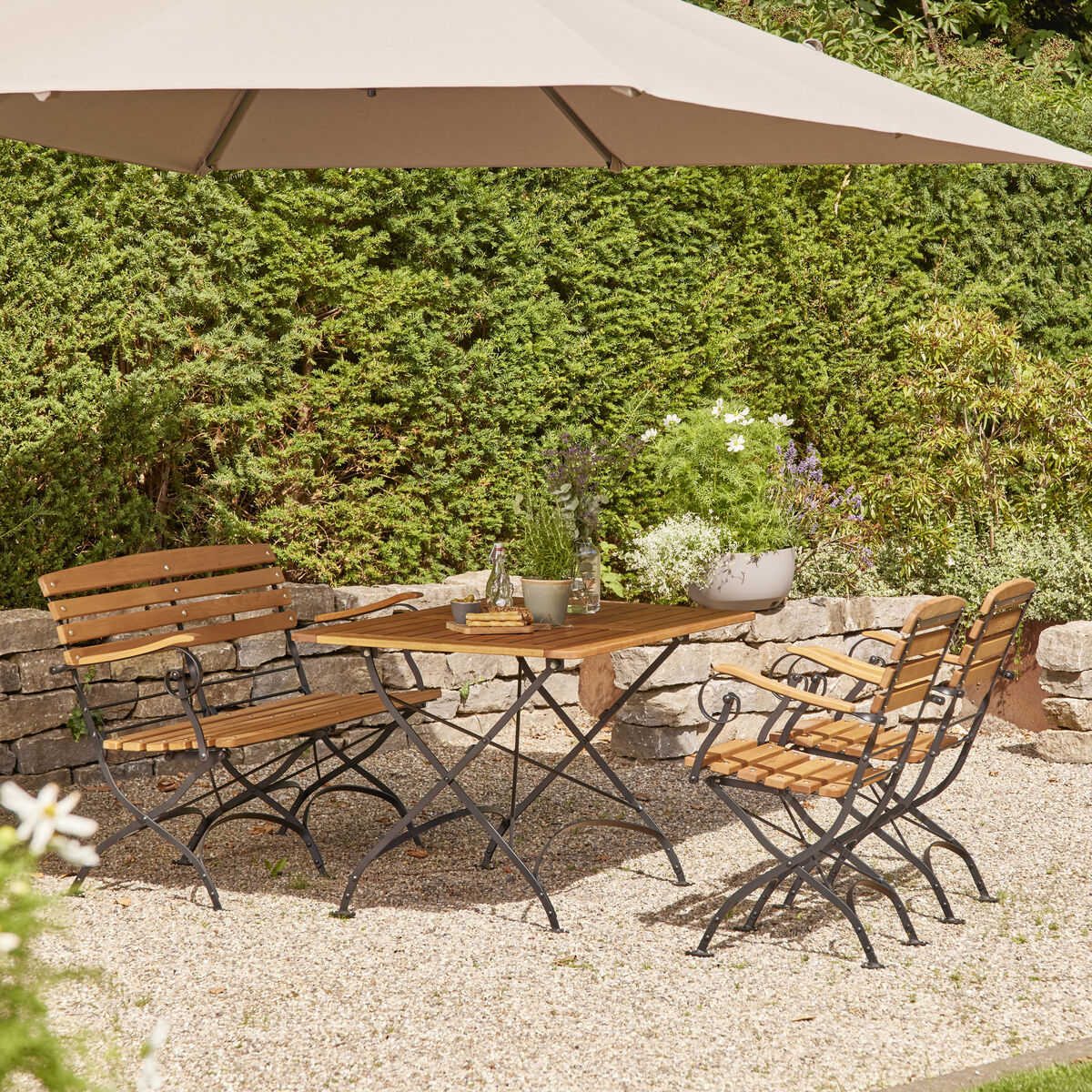 Ein Klapptisch »Peru III« aus Holz und Metall, 120x80cm groß, steht auf einer Terrasse im Garten. Er ist von vier passenden Stühlen umgeben und unter einem großen Sonnenschirm platziert. Die Umgebung ist grün und sieht gepflegt aus.