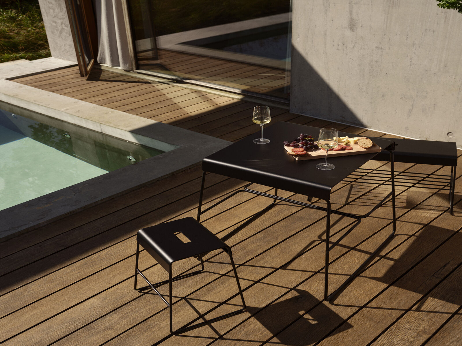 Eine moderne Outdoor-Bank mit Ablage auf einer Holzterrasse neben einem kleinen Pool. Die Szene zeigt ein minimalistisches Design mit einem kleinen Tisch, zwei Gläsern Wein und einer Holzplatte mit Snacks. Ideal für entspannte Gartenmomente.