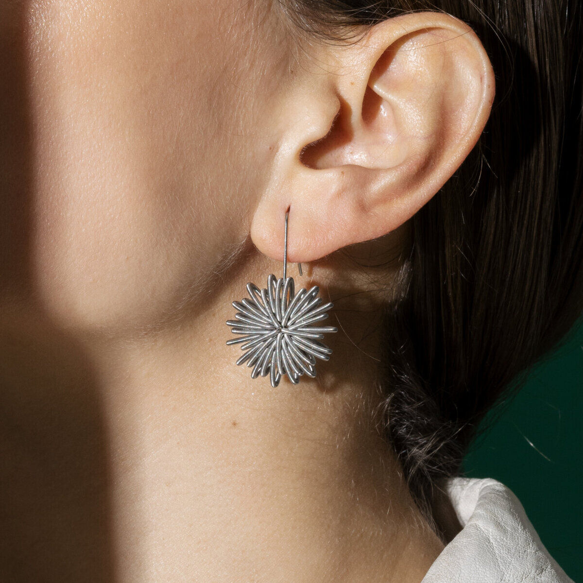 Zu sehen ist ein Ohr mit einem auffälligen, silberfarbenen Ohrring der Marke la molla. Der Ohrring „No.1 Steel“ besteht aus filigranen Edelstahlfedern in floraler Spiralform. Er sitzt leicht und elegant am Ohr. Im Hintergrund ist ein Teil eines hellen Kleidungsstücks und eine grüne Fläche erkennbar.