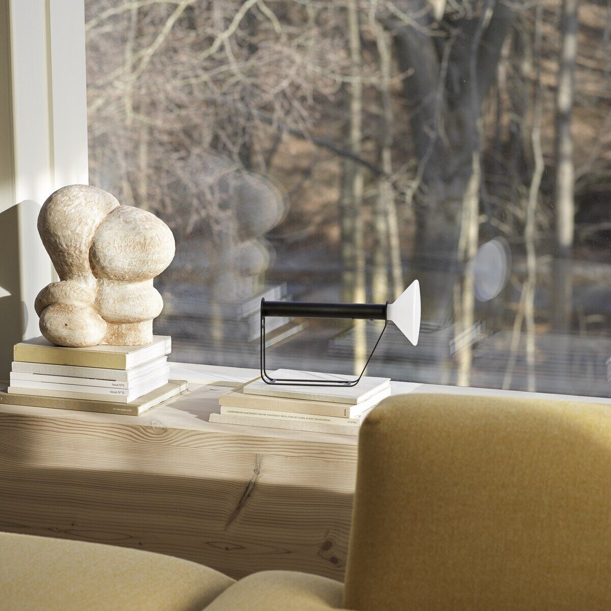 Ein modernes Wohnzimmer mit einer Tragbaren Lampe 'Piton' auf einer Holzbank neben einer kunstvollen Skulptur und mehreren Büchern vor einem großen Fenster mit Blick auf einen Wald.