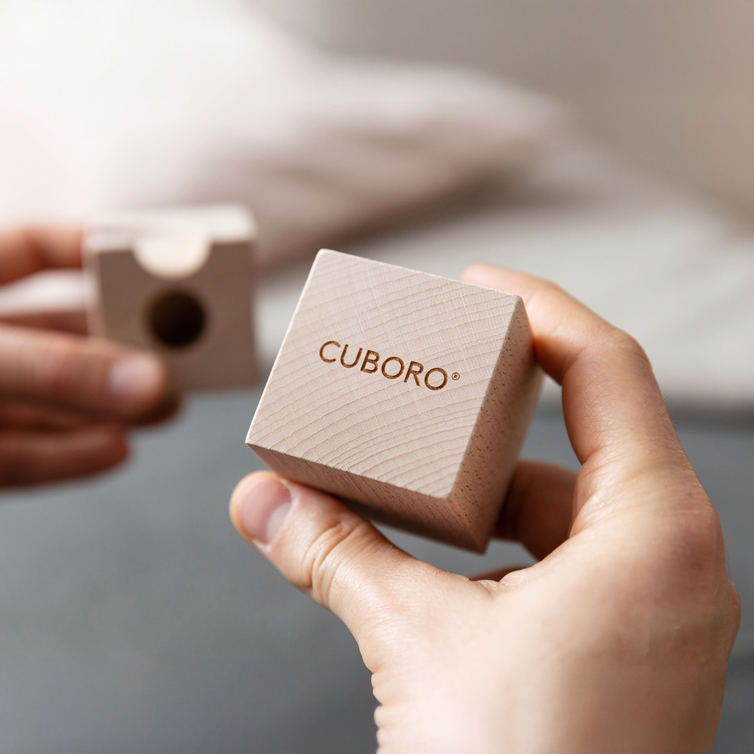 Zwei Hände halten jeweils einen Würfel aus naturbelassenem Holz aus dem CUBORO Kugelbahn Starter-Set »STANDARD«. Auf einem Würfel ist das Cuboro-Logo zu sehen, der andere Würfel hat eine eingefräste Kugelbahn-Vertiefung. Der Hintergrund ist unscharf und minimalistisch.