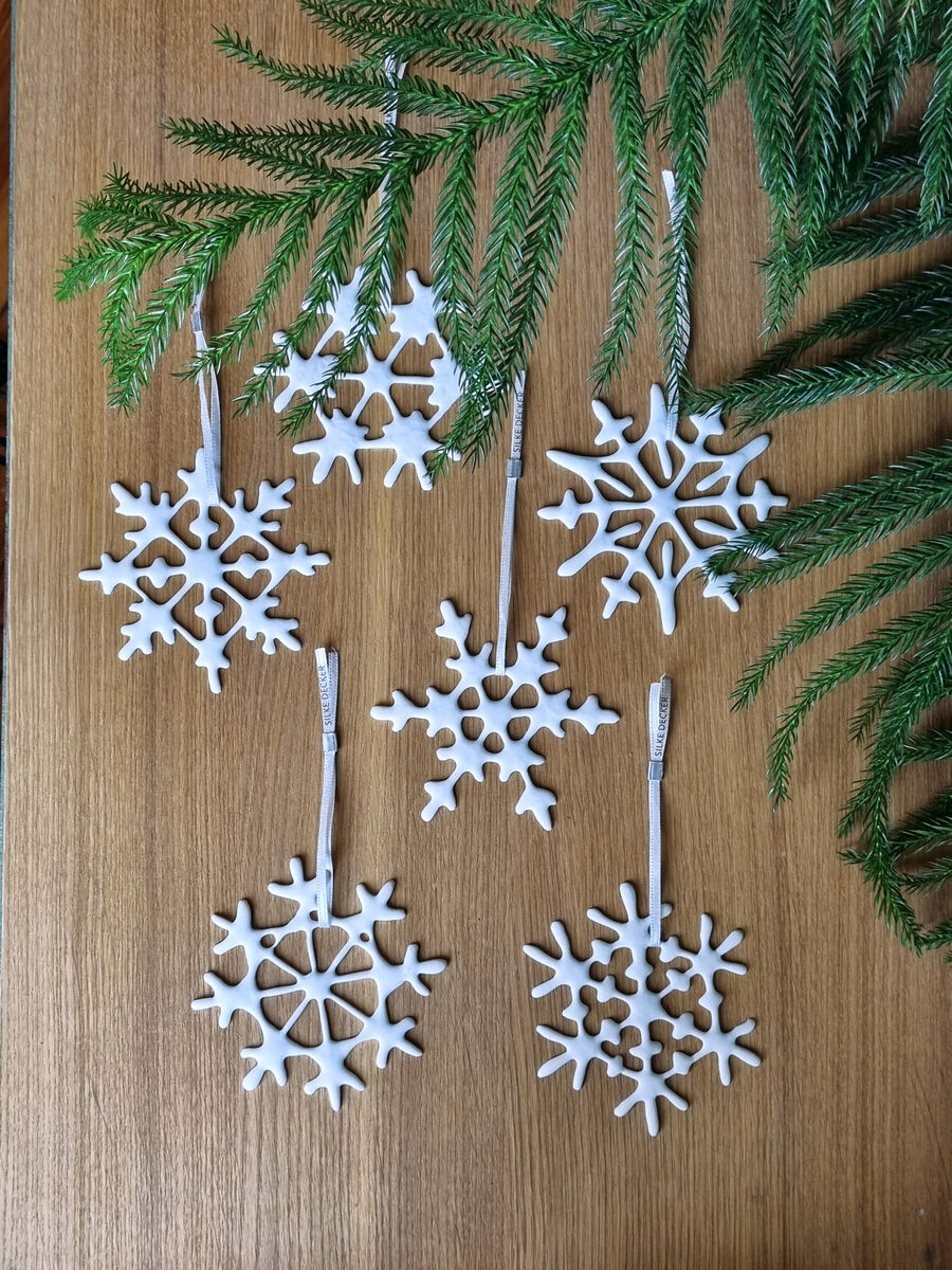 Ein Set aus sechs weißen Schneeflockenornamenten liegt auf einer braunen Holzoberfläche, umgeben von grünen Tannenzweigen. Die Schneekristalle sind aus einem dekorativen Material gefertigt und haben unterschiedliche Muster.