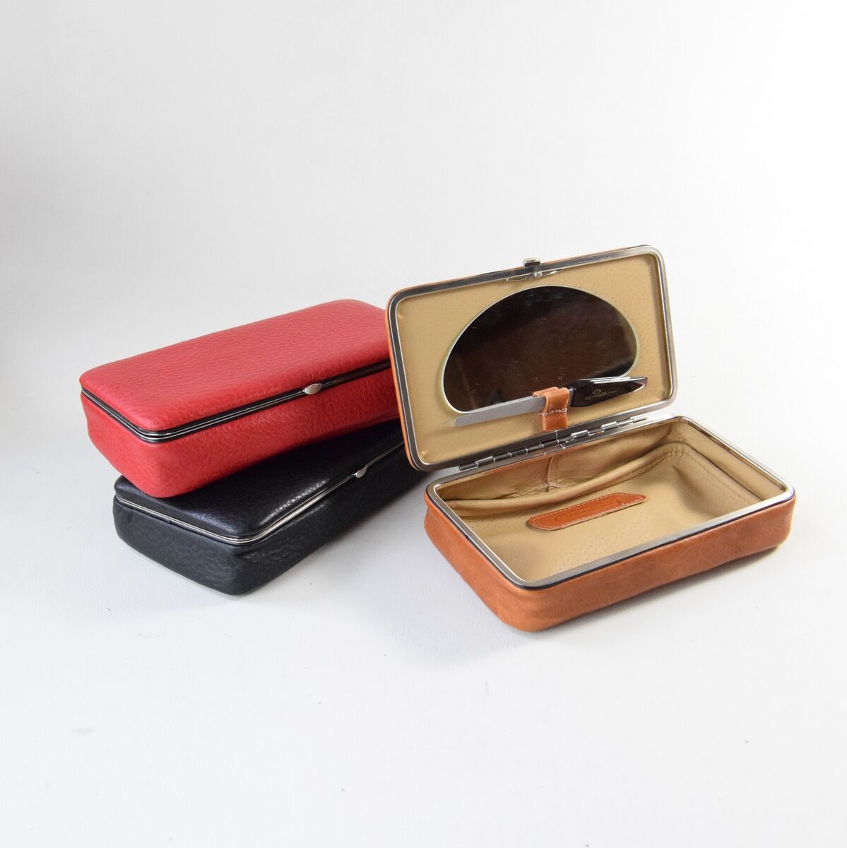 Drei Utensilienboxen aus Leder in Rot, Schwarz und Braun. Eine Box ist geöffnet und zeigt ein beigefarbenes Inneres und einen Spiegeldeckel. Die Hintergrundfarbe ist neutral.