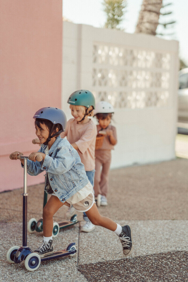 Drei Kinder fahren auf ihren Scootern im Freien. Ein Mädchen in einer Jeansjacke und einem Helm fährt vorneweg, während zwei weitere Kinder mit Helmen und Rollern hinter ihr herfahren. Der Hintergrund zeigt eine Mauer und einen Baum, es scheint ein sonniger Tag zu sein.