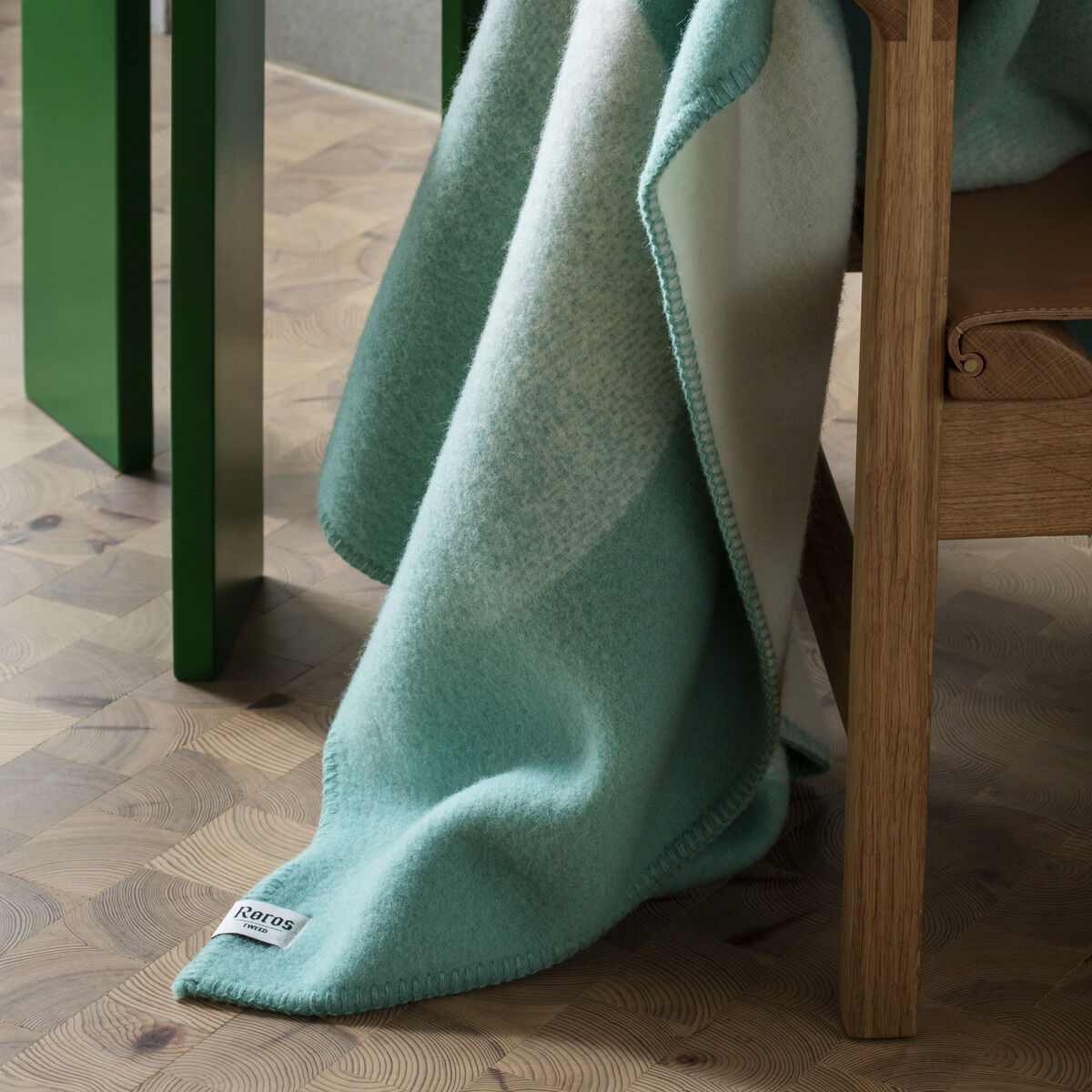 Ein weicher, grüner und weißer Lammwoll-Deckenüberwurf liegt auf einem hölzernen Sessel neben einem grünen Beistelltisch mit einer Tasse und einer Tischlampe.