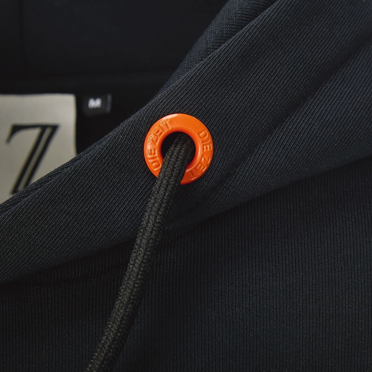 Nahaufnahme eines schwarzen Hoodies aus der ZEIT-Kollektion »Verbrechen«. Der Hoodie besitzt Kordeln mit einem orangefarbenen Ring, auf dem 'DIE ZEIT' steht. Ein Label mit einem 'Z' ist ebenfalls sichtbar.