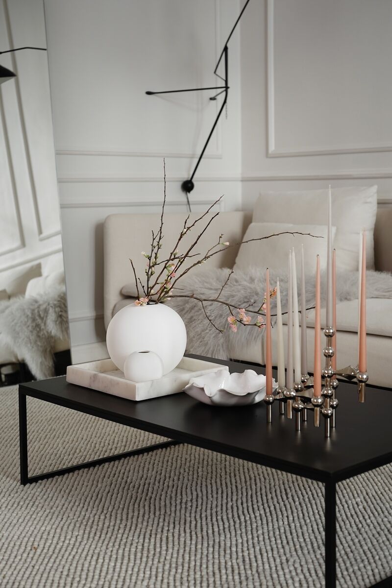 Ein moderner Kerzenständer aus Chrom steht auf einem schwarzen Couchtisch in einem elegant eingerichteten Wohnzimmer, umgeben von minimalistischen Dekorationselementen in neutralen Farben.