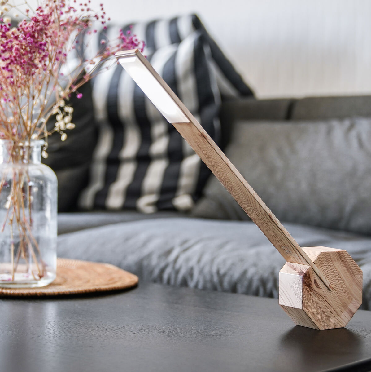 Ein Wohnzimmer mit einer modernen Octagon Tischlampe »One« aus Holz auf einem schwarzen Tisch. Daneben steht eine Vase mit rosa und weißen Trockenblumen. Im Hintergrund ist ein graues Sofa mit gestreiften Kissen sichtbar.