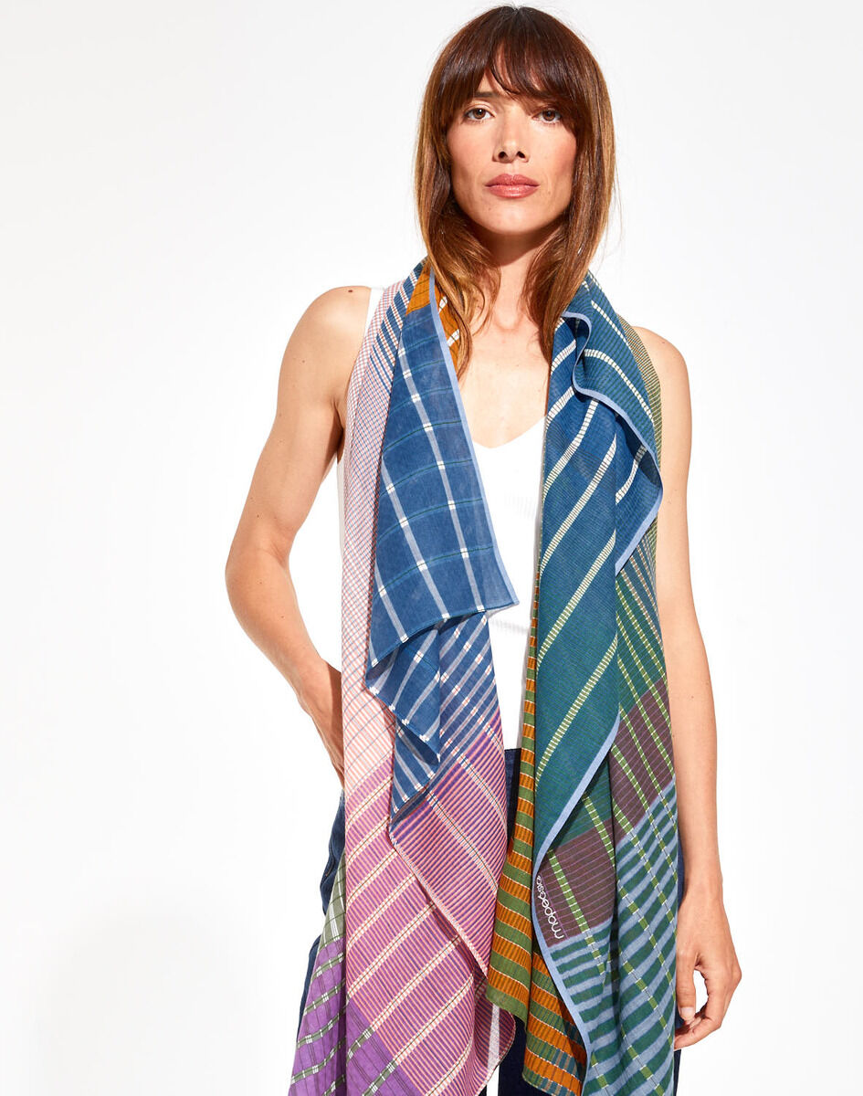 Frau mit einem bunten Baumwolltuch in Streifenmuster, das in Blau, Grün, Pink und Braun gehalten ist; die Frau trägt ein weißes Oberteil, und das Tuch wirkt leicht und elegant.