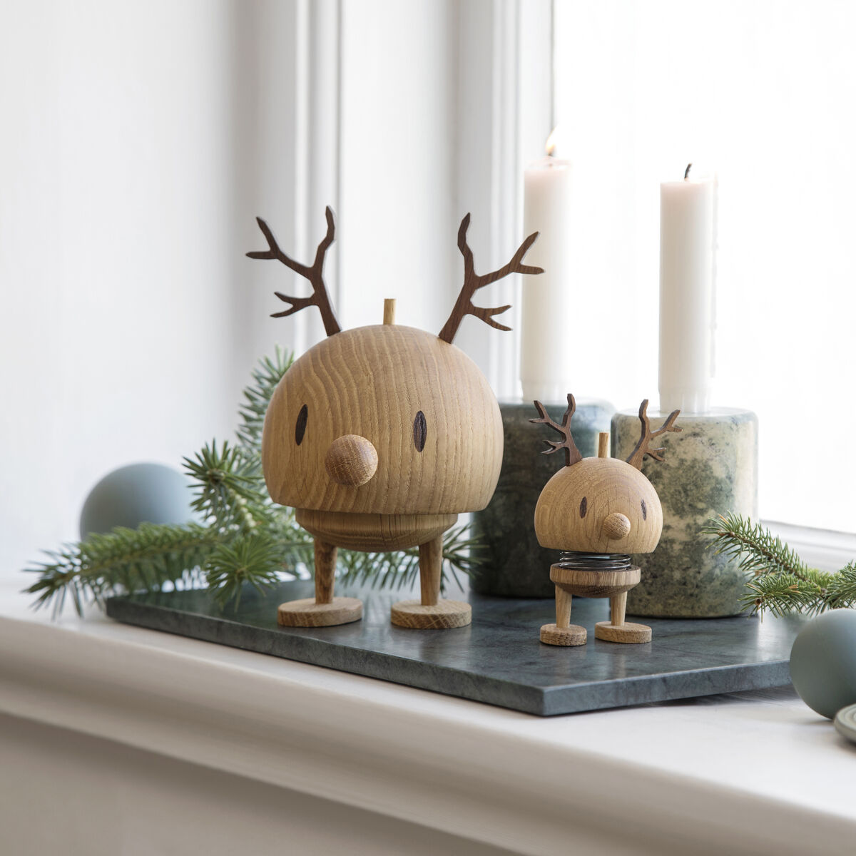 Zwei Hoptimist Rentier Bumble Figuren aus Eiche stehen auf einer Fensterbank, umgeben von grünen Tannenzweigen, hellblauen Kugeln und weißen Kerzen in verschiedenen Haltern. Die Szene wirkt modern, minimalistisch und eignet sich als festliche Weihnachtsdekoration.