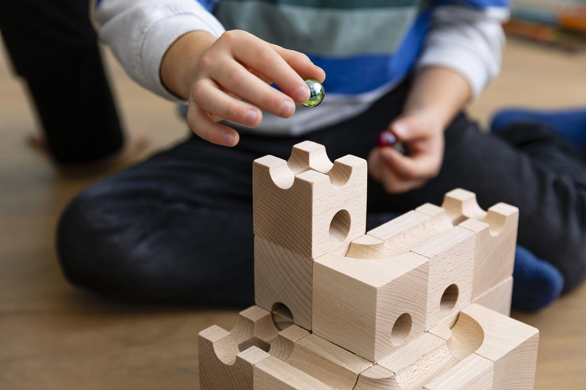 Ein Kind spielt mit einer Kugelbahn Starter-Set »STANDARD«. Es ist aus Holz gefertigt und enthält mehrere Bausteine, bei denen Murmeln durch verschiedene Bahnen rollen können. Die Hände des Kindes halten eine Murmel über der Bahn.
