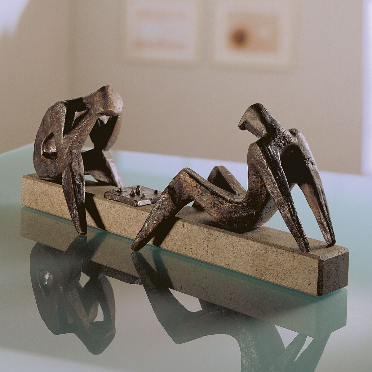 Detaillierte Bronzeskulptur von Sepp Mastaller mit zwei abstrahierten, stilisierten Personen, die Schach spielen. Die Figuren sitzen auf einem länglichen Stein-Sockel, das Schachbrett liegt zwischen ihnen. Das Kunstwerk steht auf einer reflektierenden Glasoberfläche in modernem, hellen Raum.