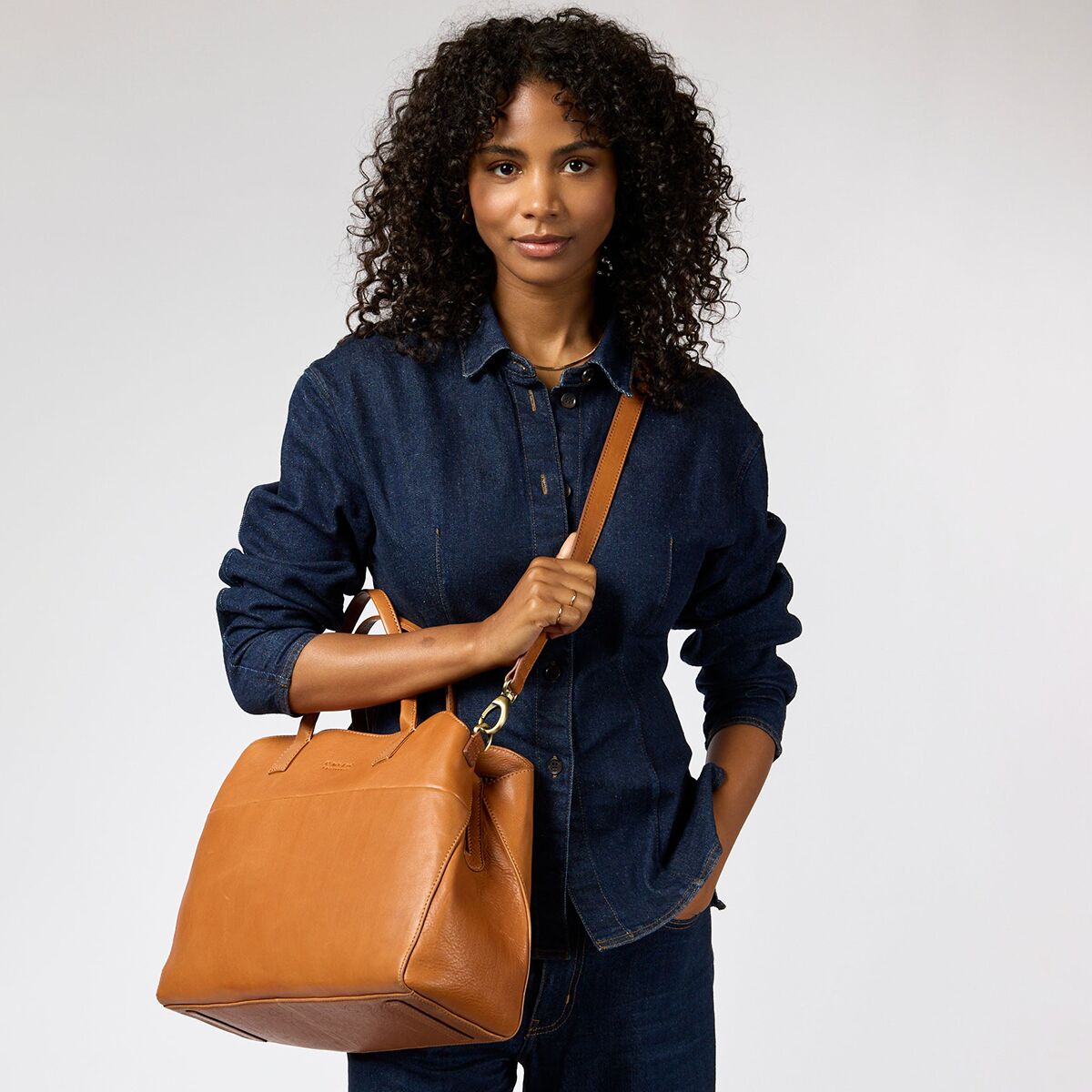 Eine Frau trägt eine mittelgroße, hellbraune Schultertasche aus Narbenleder von O My Bag über der Schulter. Sie trägt ein dunkelblaues Jeanshemd und hält die Tasche mit einer Hand, während der längenverstellbare Schulterriemen locker fällt. Die Tasche hat stabile Griffe, goldene Beschläge und eine schlichte, elegante Form.