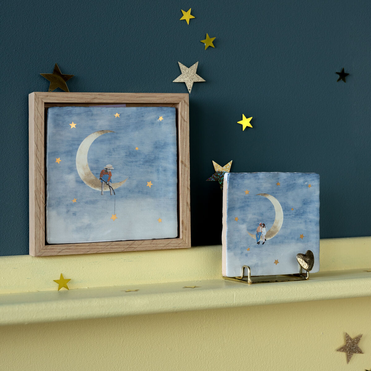 Keramikfliese in einem Holzrahmen zeigt ein Bild von einem Mann, der auf der Mondsichel sitzt, umgeben von gelben Sternen auf einem blauen Hintergrund. Mehrere sternförmige Dekorationen umgeben das Bild.