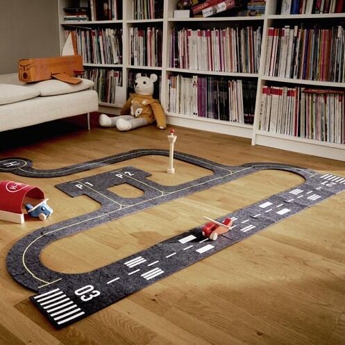 Das Bild zeigt ein Spielset »gokiAir« mit Flughafen-Thema, ausgelegt auf einem Holzfußboden in einem hellen Kinderzimmer. Zu sehen sind eine schwarze Start- und Landebahn mit Aufdrucken, kleine bunte Holzflugzeuge, ein Tower und ein Hangar. Im Hintergrund steht ein Kuscheltier auf einem Sofa vor einem Bücherregal. Das Set ist geeignet für fantasievolles Spielen und ideal für Kinder.