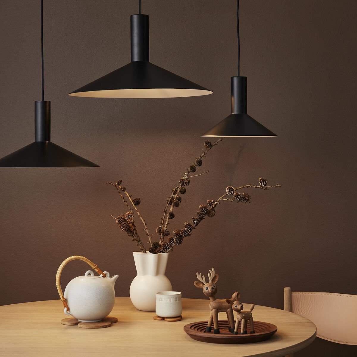 Auf einem runden Holztisch stehen eine schlichte Teekanne, eine kleine Tasse, eine weiße Vase mit Zweigen und ein rundes Tablett mit einer stilisierten Holzfigur »Spirit« in Form eines Rehs. Im Hintergrund hängen drei schwarze Pendelleuchten vor einer braunen Wand. Die Szene wirkt minimalistisch und warm.