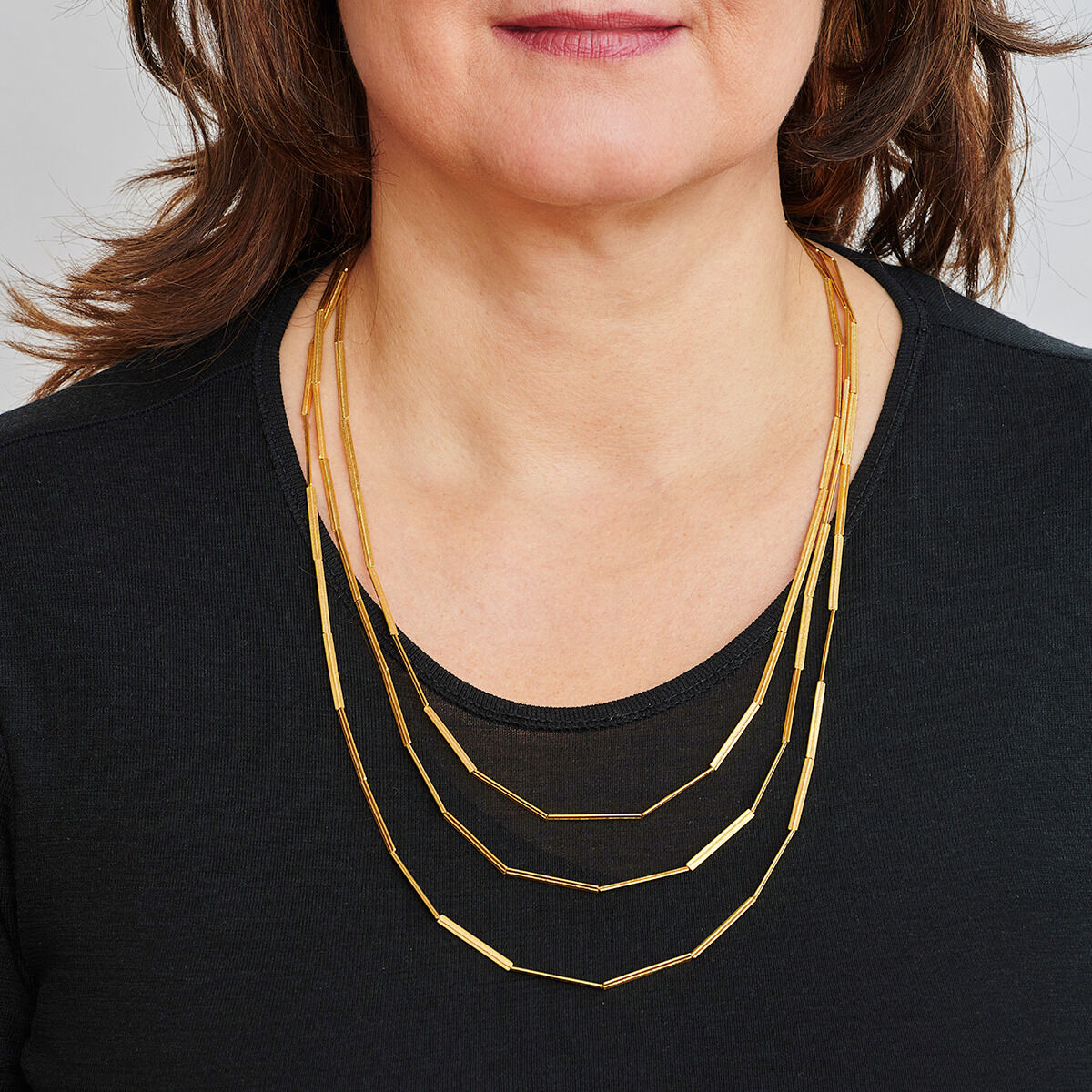 Nahaufnahme einer Frau, die eine goldplattierte, mehrreihige Kette namens »Streifen« trägt. Die Kette besteht aus drei dünnen, goldenen Streifen, die um ihren Hals liegen. Sie trägt ein schwarzes Oberteil.