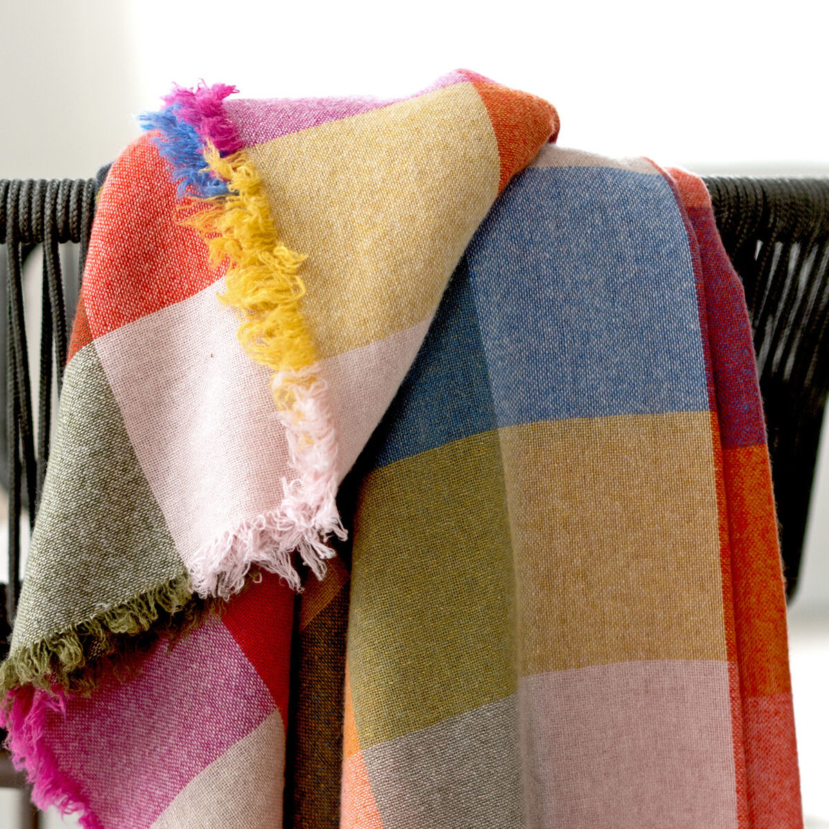 Eine farbenfrohe Merino-Kaschmir-Decke »St. Tropez« mit großem Karomuster und Fransen liegt dekorativ über der Lehne eines modernen schwarzen Stuhls. Die Decke zeigt leuchtende Farbfelder in Rosa, Blau, Gelb, Beige und mehr. Der helle, minimalistische Raum wirkt ruhig und einladend.