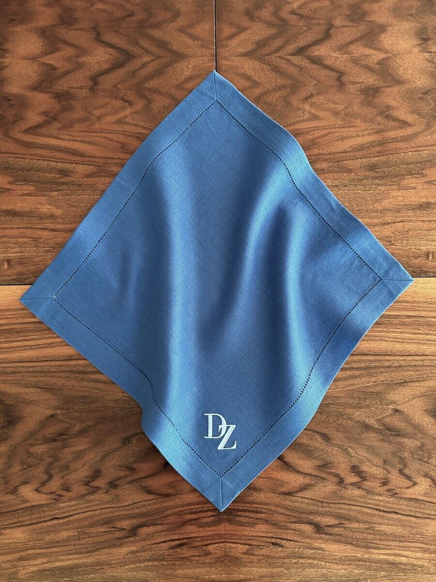 Blaue Leinenserviette mit Monogramm 'DZ' auf einem dunkel gemaserten Holztisch liegend. Die Serviette ist sorgfältig gefaltet und zeigt eine präzise genähte Kante.