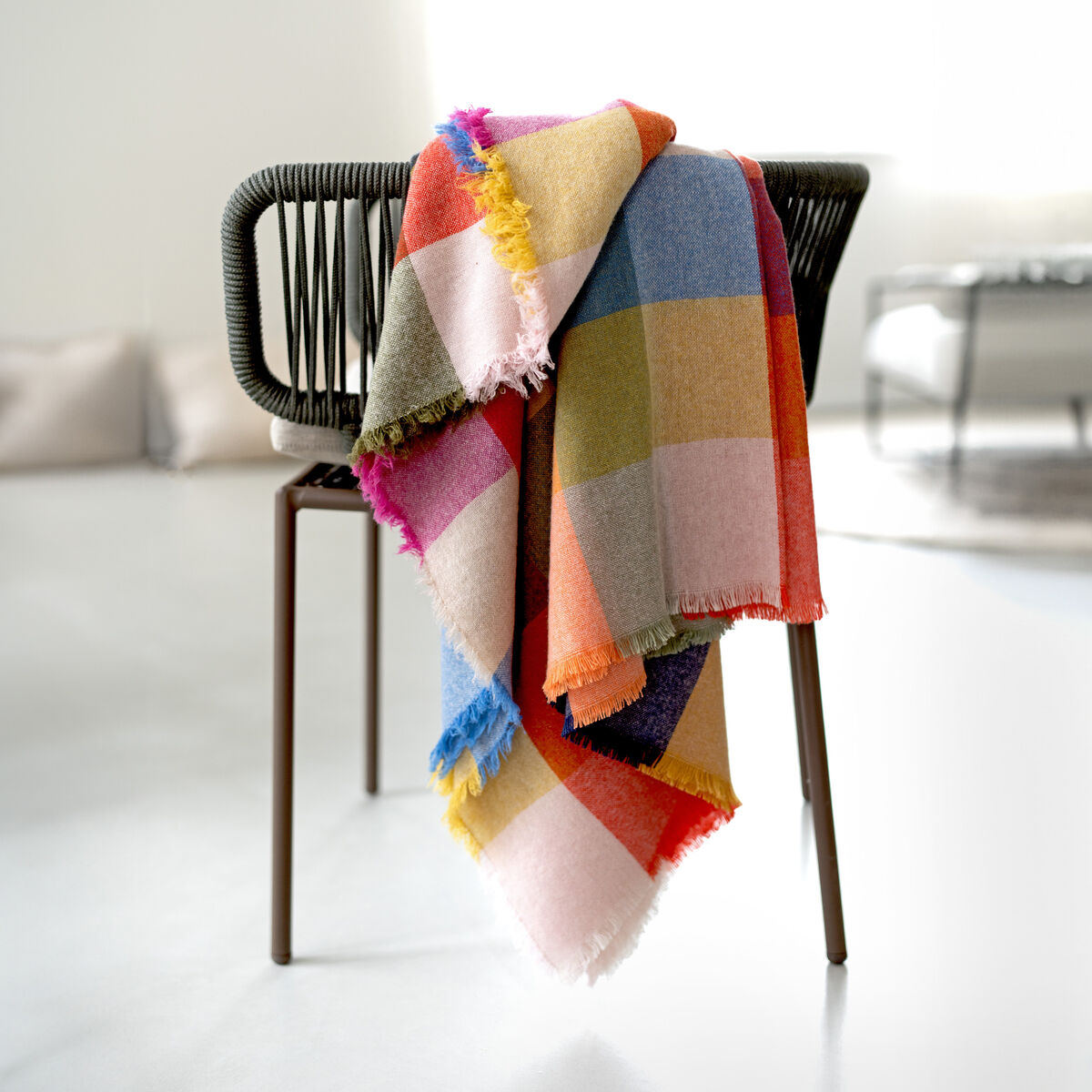 Eine farbenfrohe Merino-Kaschmir-Decke »St. Tropez« mit großem Karomuster und Fransen liegt dekorativ über der Lehne eines modernen schwarzen Stuhls. Die Decke zeigt leuchtende Farbfelder in Rosa, Blau, Gelb, Beige und mehr. Der helle, minimalistische Raum wirkt ruhig und einladend.