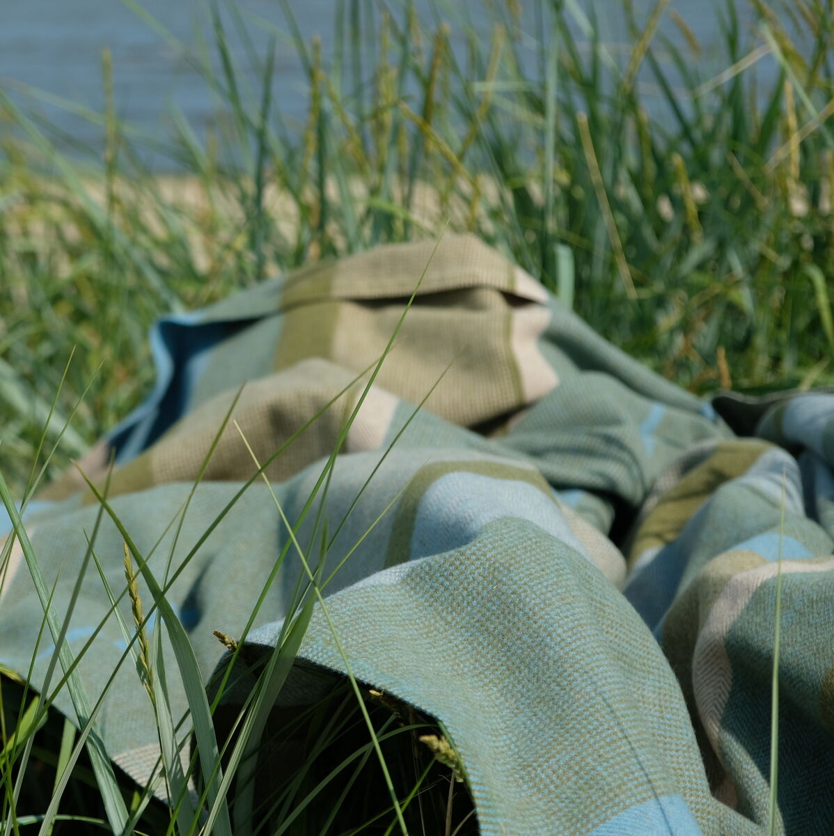 Eine Decke mit grün, beige und hellblauem Muster liegt auf einer Wiese, im Hintergrund ist Wasser zu sehen.