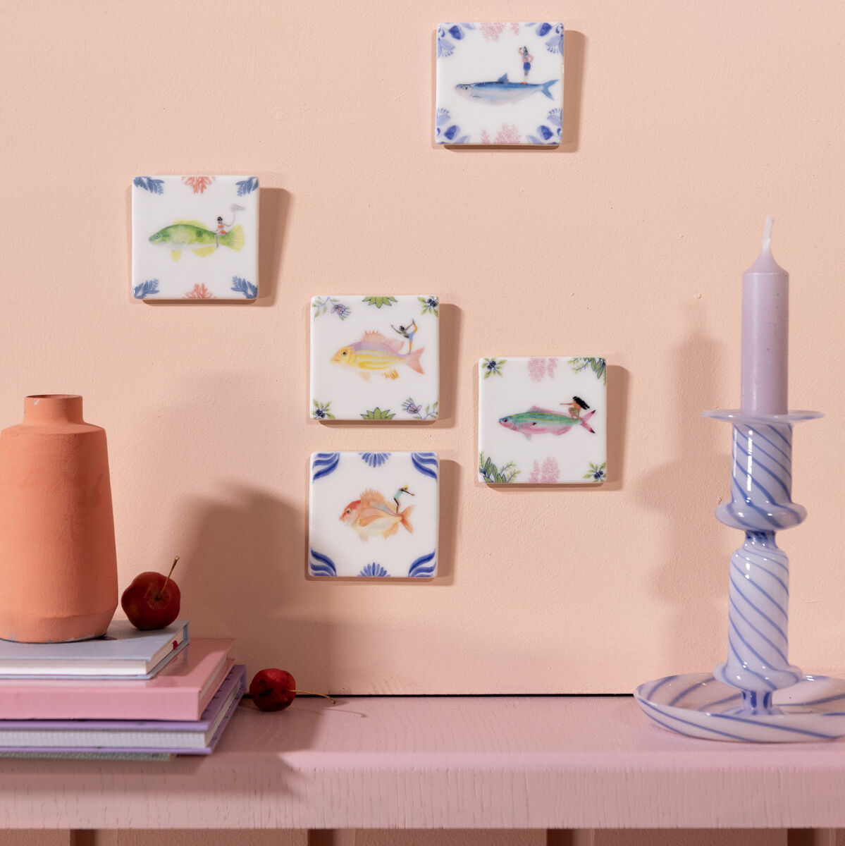 Keramikfliesen auf einer pastellrosa Wand, arrangiert in einem Muster. Die Fliesen zeigen handgemalte Meerestiere und Naturmotive. Eine graue Kerze auf einem hellblauen Kerzenhalter und eine Terrakottavase stehen auf einem Regal darunter.