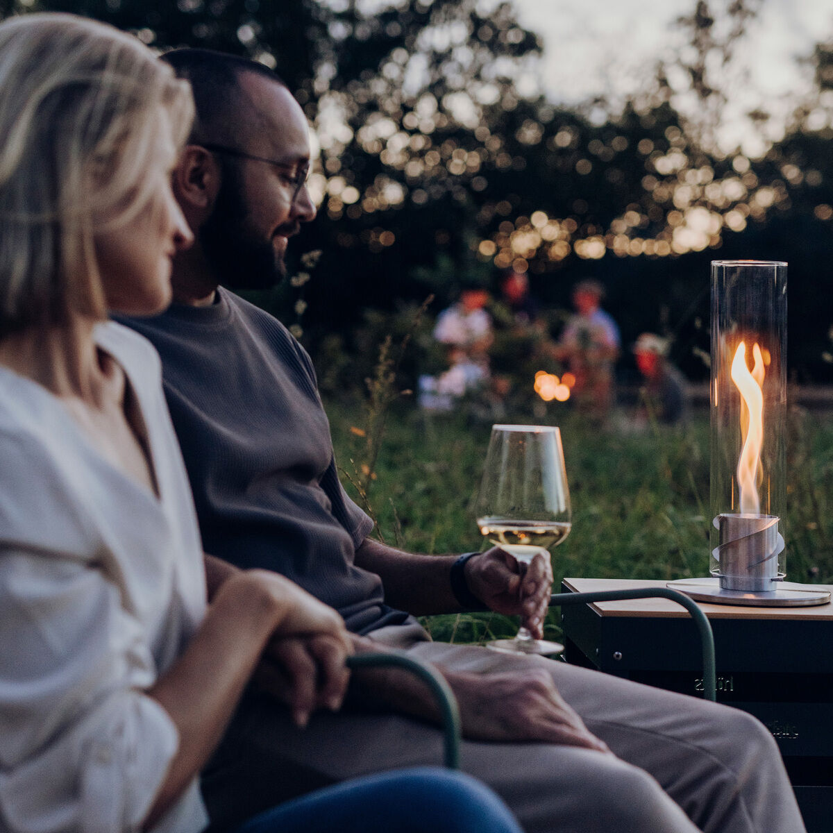Ein Paar sitzt bei Dämmerung auf Rasenstühlen im Garten. Auf einem Tisch zwischen ihnen brennt ein Tischfeuer »SPIN 900«. Der Mann hält ein Weinglas. Im Hintergrund sind Bäume und weitere Personen zu sehen.