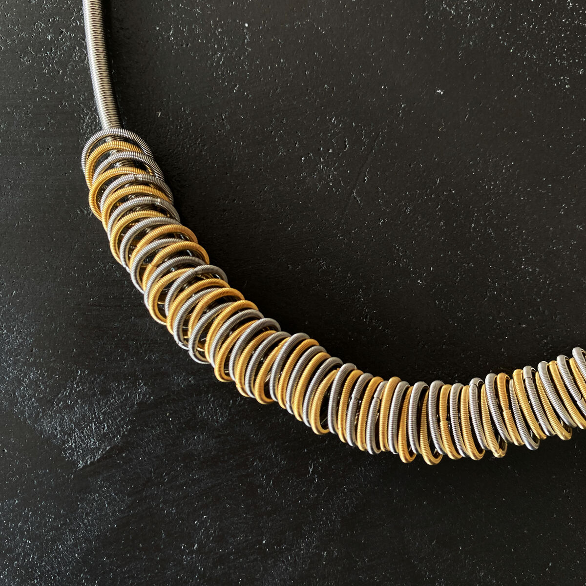 ZEIT-Sonderedition Collier »ZEIT«, bestehend aus einer Reihe goldener und silberner Metallringe, die auf einem flexiblen Halsband in einer Wellenform angeordnet sind, vor einem schwarzen Hintergrund.