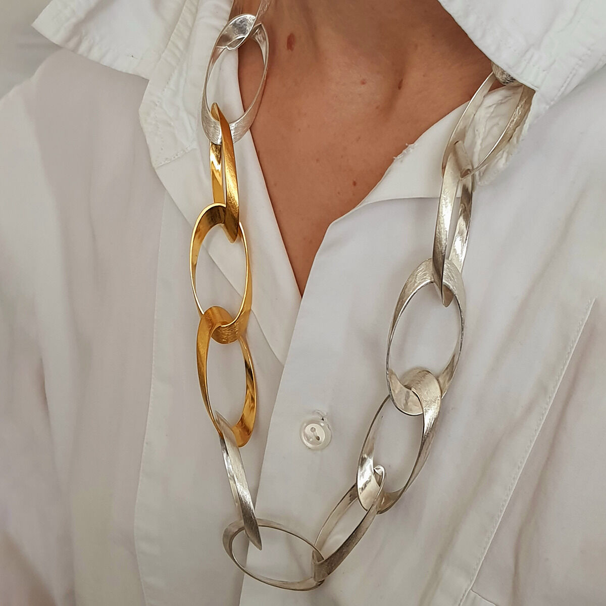 Eine Person trägt das HML-Collier »Standing Ovations« von HML Berlin aus 925er Silber und Feingoldplattierung. Die Kette hat große, ovale Glieder in Silber und Gold, die abwechselnd angeordnet sind. Der Hintergrund ist ein weißes Hemd, Szene wirkt elegant und modern.