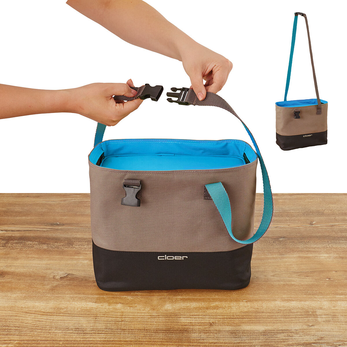 Das Bild zeigt eine Lunch-Tasche in Grau und Schwarz mit blauen Akzenten auf einem Holztisch. Zwei Hände verschließen die Tasche mit einem Steckverschluss. Im Hintergrund ist die geöffnete Tasche mit einem blauen Innenfutter und einem stabilen blauen Trageriemen zu sehen.