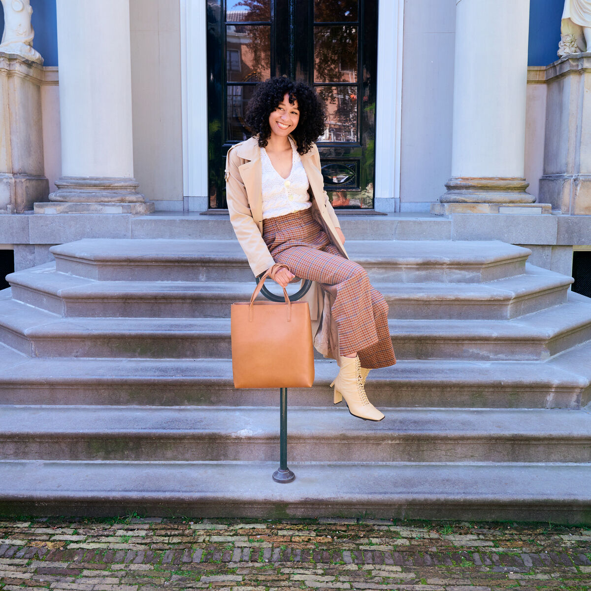 Eine Frau sitzt auf einer Treppe vor einem Gebäude und hält eine braune Ledertasche »Georgia«. Sie trägt eine beige Jacke, einen rötlich-braunen Rock, einen weißen Pullover und beige Stiefel.