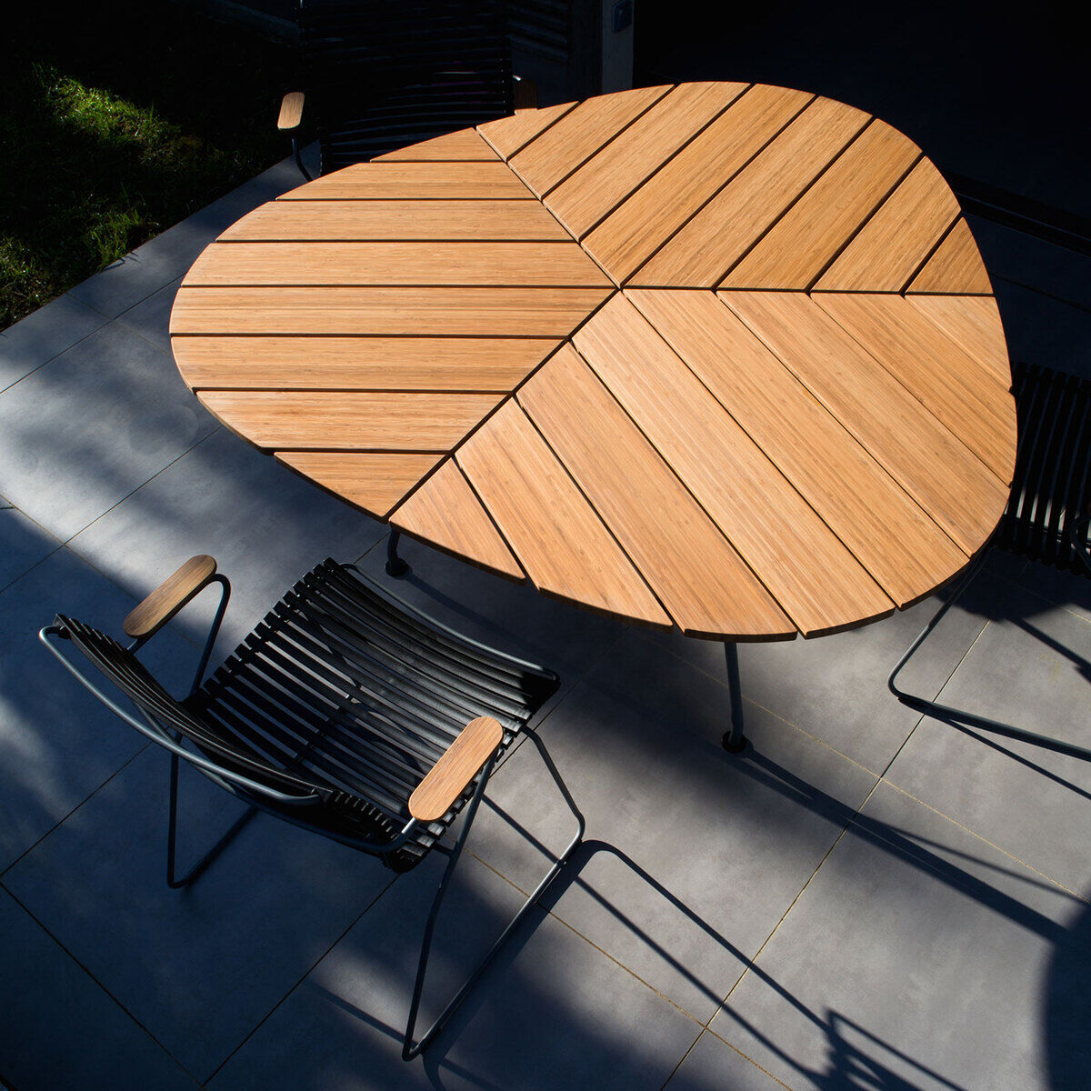 Moderner Gartentisch namens 'LEAF' mit einzigartigem Holzdesign, umgeben von stilvollen schwarzen Gartenstühlen, auf einer hellgrauen Terrasse.
