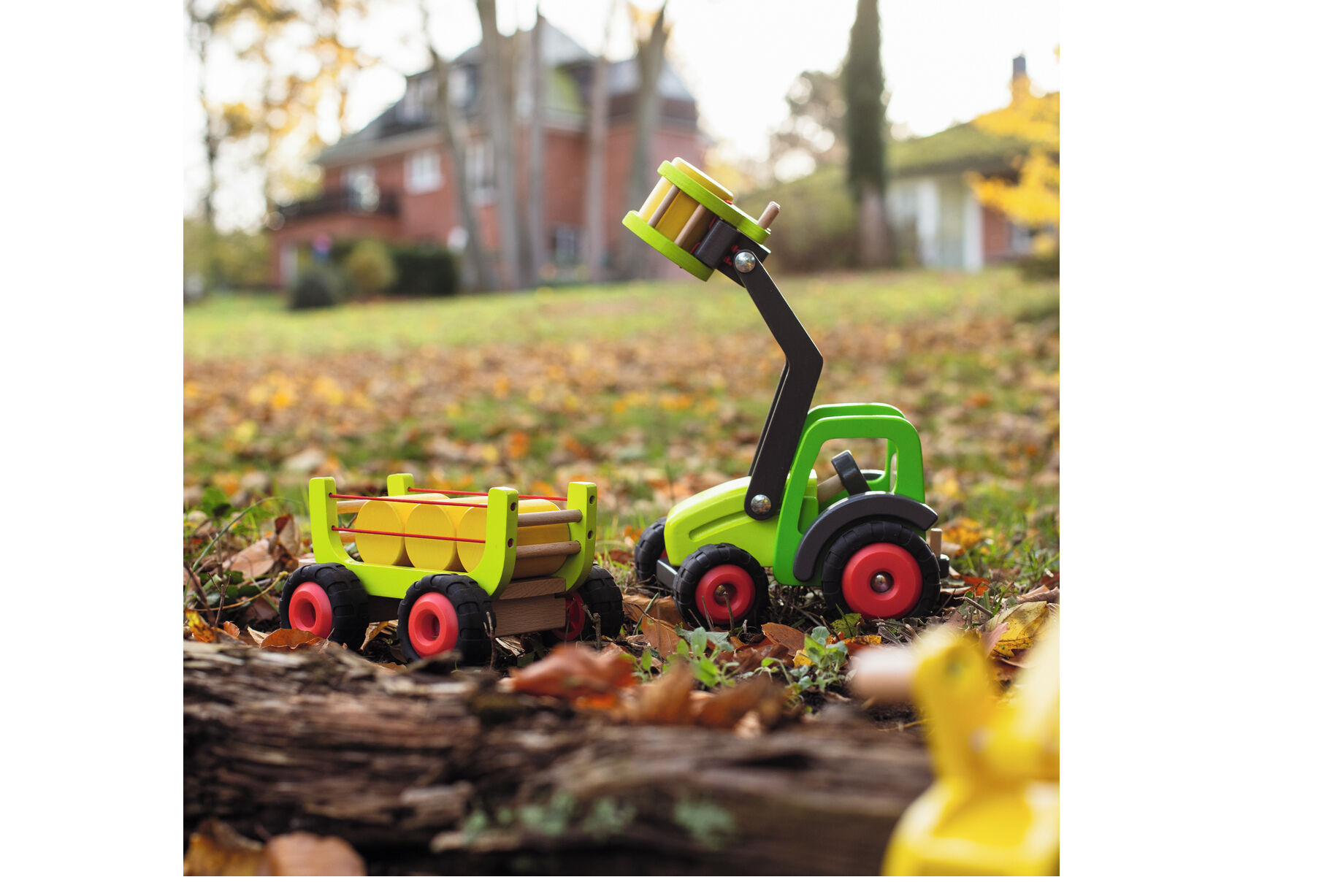 Ein Holz-Frontlader mit Heuwagen steht auf einem mit Herbstlaub bedeckten Garten. Das robuste und bunte Holzspielzeug mit beweglichem Arm und kleinen Holzstämmen im Anhänger ist ideal für Kinder. Im Hintergrund sind Bäume, Laub und ein Haus in herbstlicher Atmosphäre zu sehen.