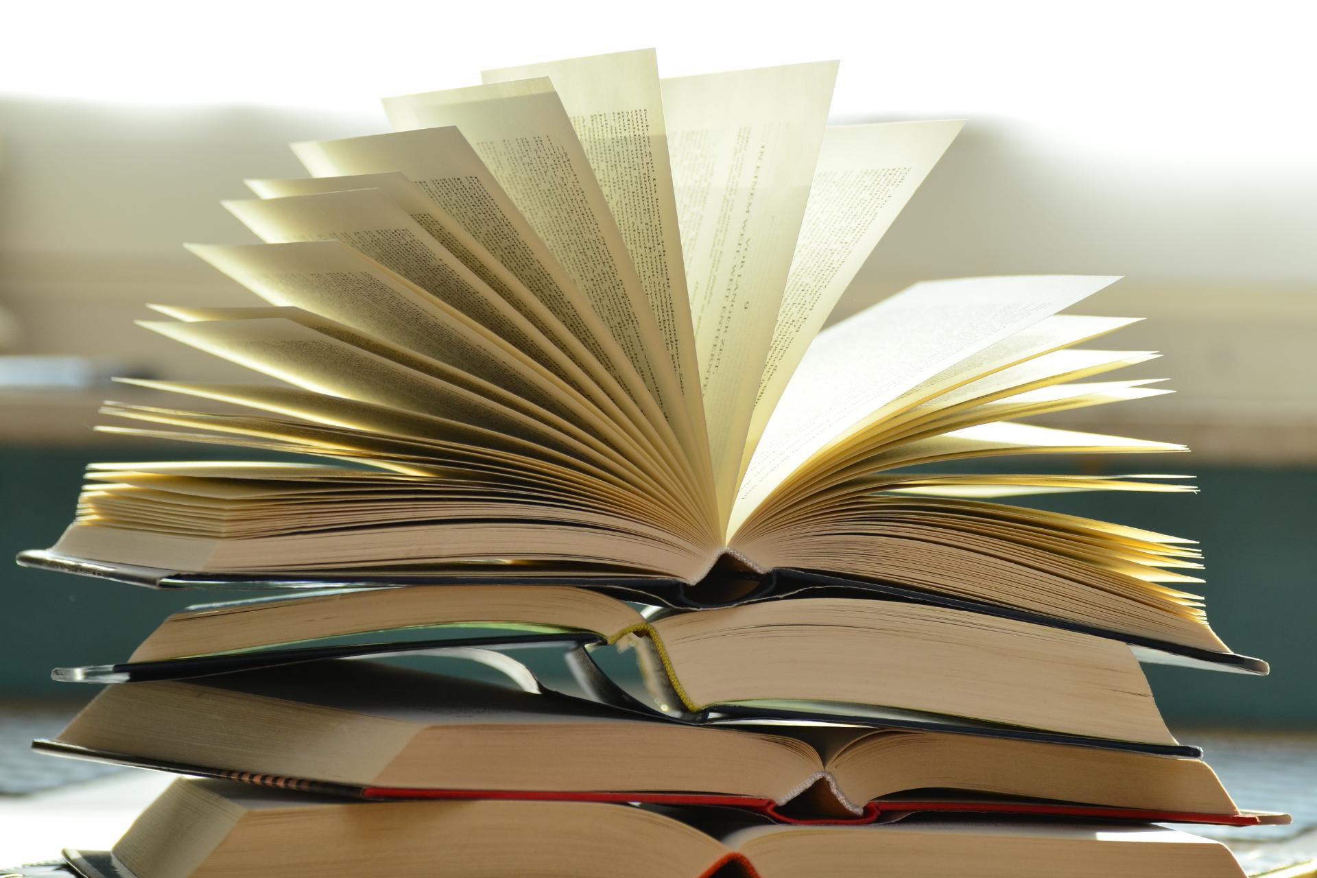Link zur die besten Bücher 2025. Mehrere aufgeschlagene Bücher liegen übereinander, ihre Seiten bilden auffächernde Muster. Die Seiten sind cremefarben bis weiß, die Buchrücken und -umschläge zeigen gedeckte Farben wie Rot, Grün, Beige und Blau. Der Hintergrund ist weiß und hell ausgeleuchtet.