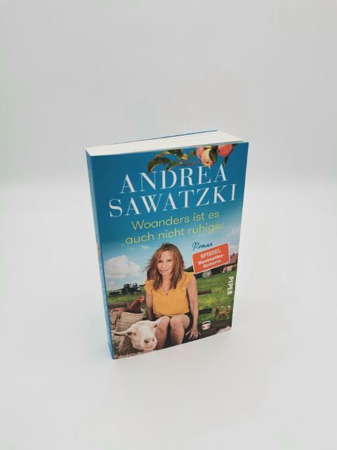 Das Buch 'Andrea Sawatzki: Woanders ist es auch nicht ruhiger' zeigt auf dem Cover ein lächelndes Porträt von Andrea Sawatzki auf einem Bauernhof. Sie sitzt neben einem weißen Schwein vor einem blauen Himmel mit einem Apfelbaum.