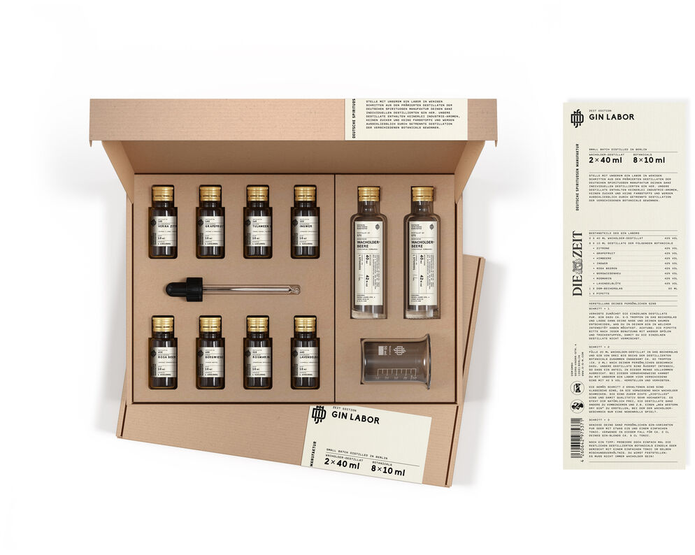 ZEIT-Sonderedition DSM »Gin Labor« ist ein Set, das mehrere kleine Flaschen Gin, zwei größere Flaschen, eine Pipette, ein kleiner Messbecher und eine Karteikarte, alles in einer braunen Box enthält. Auf der rechten Seite des Bildes befindet sich die Rückseite der Box, die detaillierte Inhalte und Anweisungen zeigt.