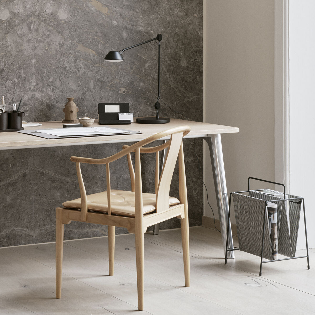 Ein modernes Arbeitszimmer mit einer minimalistischen Tischlampe AQ01 von Fritz Hansen. Die Lampe steht auf einem Holzschreibtisch zusammen mit Büchern und Büromaterial. Der Raum hat helle Holzböden, eine graue Wand und einen Holzstuhl.