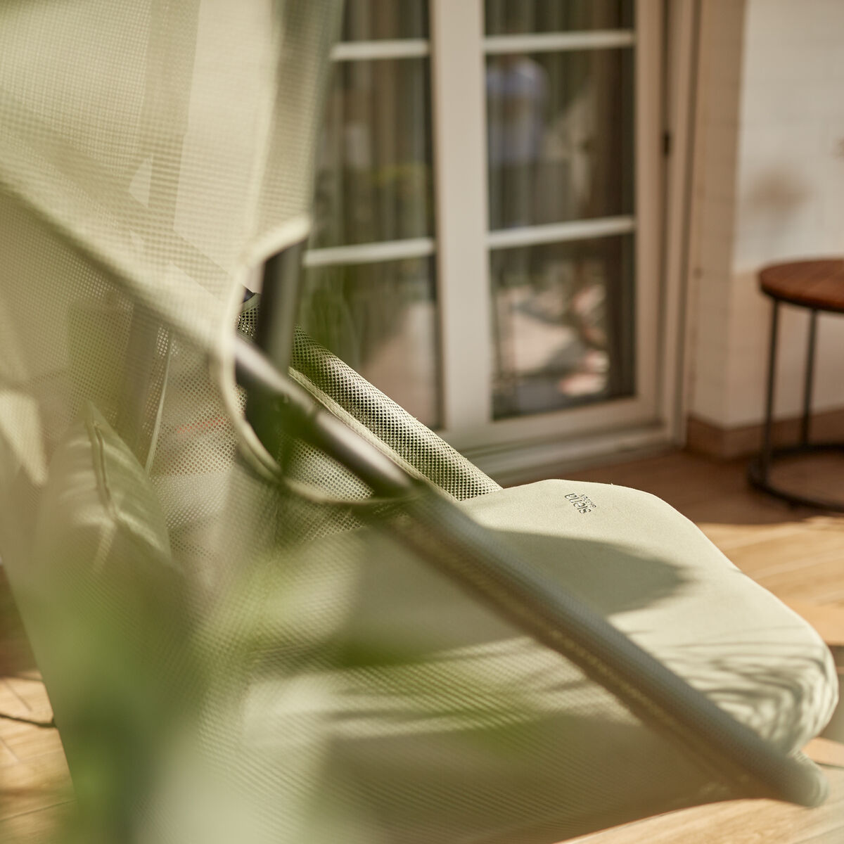 Der Hängesessel »Yobaya« von Siena Garden in hellgrünem Air Mesh Gewebe steht auf einer Terrasse nahe einer weißen Doppeltür. Im Vordergrund sind stilisierte Pflanzen zu sehen, ein runder Beistelltisch mit Vase und Topfpflanze komplettieren das harmonische Gartenambiente.