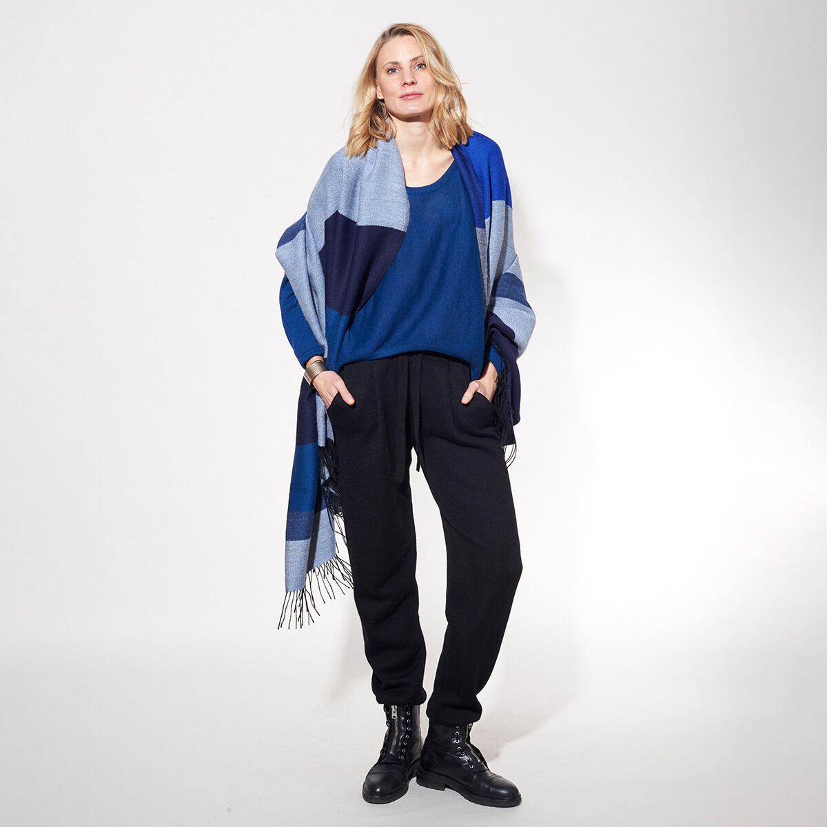 Frau trägt eine elegante Cape-Jubiläumsedition »Shades of Blue«, bestehend aus verschiedenen Blau-Schattierungen. Der Cape-Schal ist locker um ihre Schultern drapiert, und sie trägt dazu schwarze Hosen und schwarze Stiefel. Der Hintergrund ist neutral und lenkt die Aufmerksamkeit auf das Outfit.