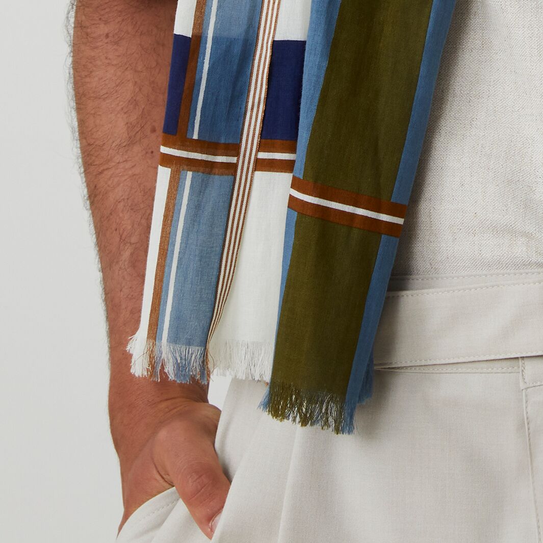 Detailansicht eines locker um den Hals gelegten Schals aus 100% Baumwolle von MOISMONT. Das Muster besteht aus breiten Längsstreifen in Blau, Grün und Weiß mit schmaleren braunen Akzentstreifen, leicht fransiger Kante und feiner Webstruktur.
