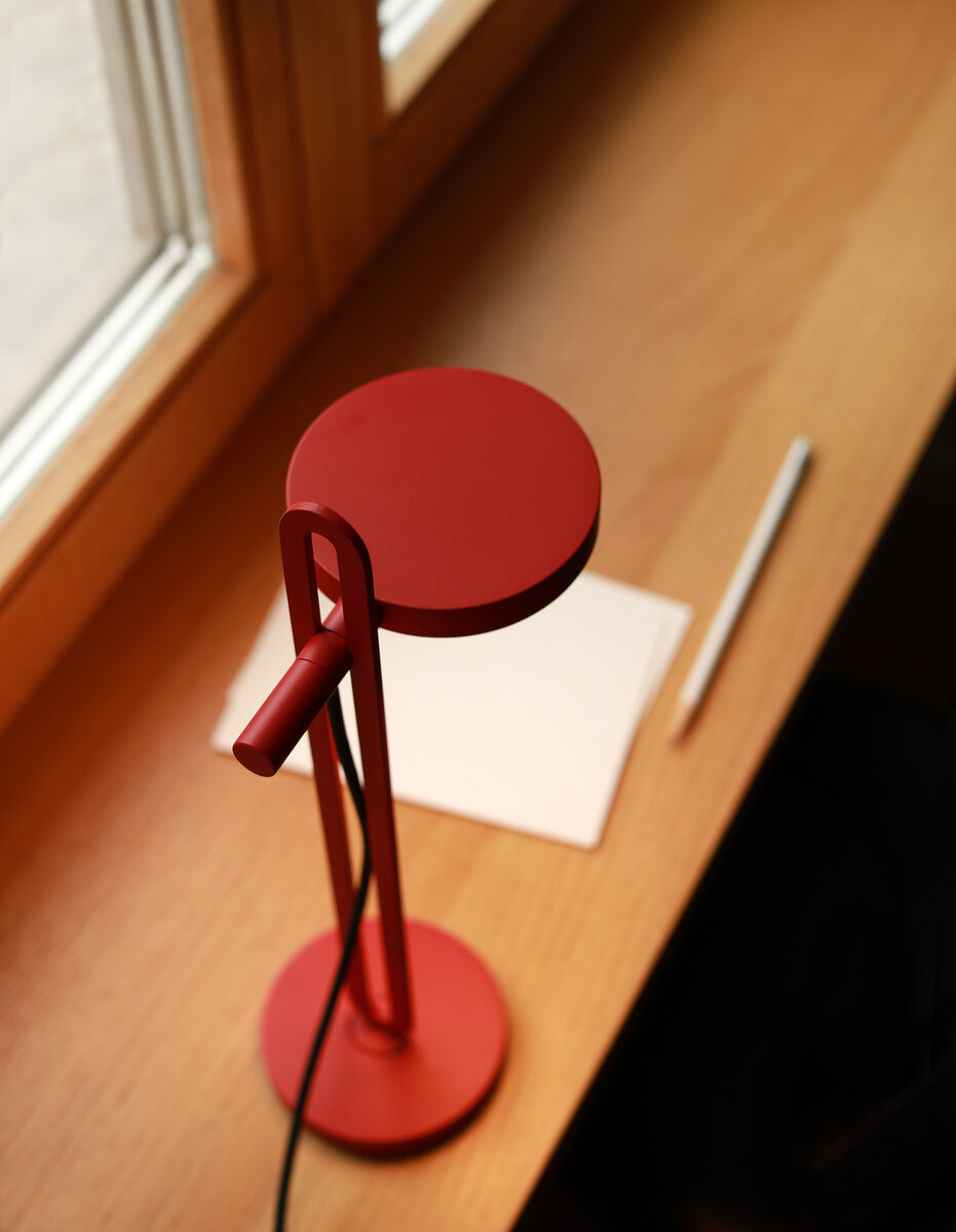 Eine rote Tischlampe mit minimalistischem Design steht auf einem hölzernen Tisch vor einem Fenster. Neben der Lampe liegt ein weißes Blatt Papier und ein Stift.