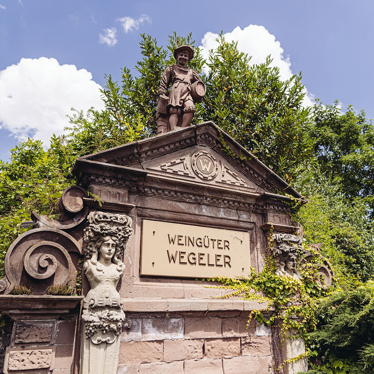 Historisches Eingangstor der Weingüter Wegeler mit detailreicher Sandsteinarchitektur, klassizistischen Säulen, dekorativen Weinranken und einer Figur am Giebel. Die Inschrift „Weingüter Wegeler“ ist gut sichtbar. Umgeben von sattem Grün und blauem Himmel.