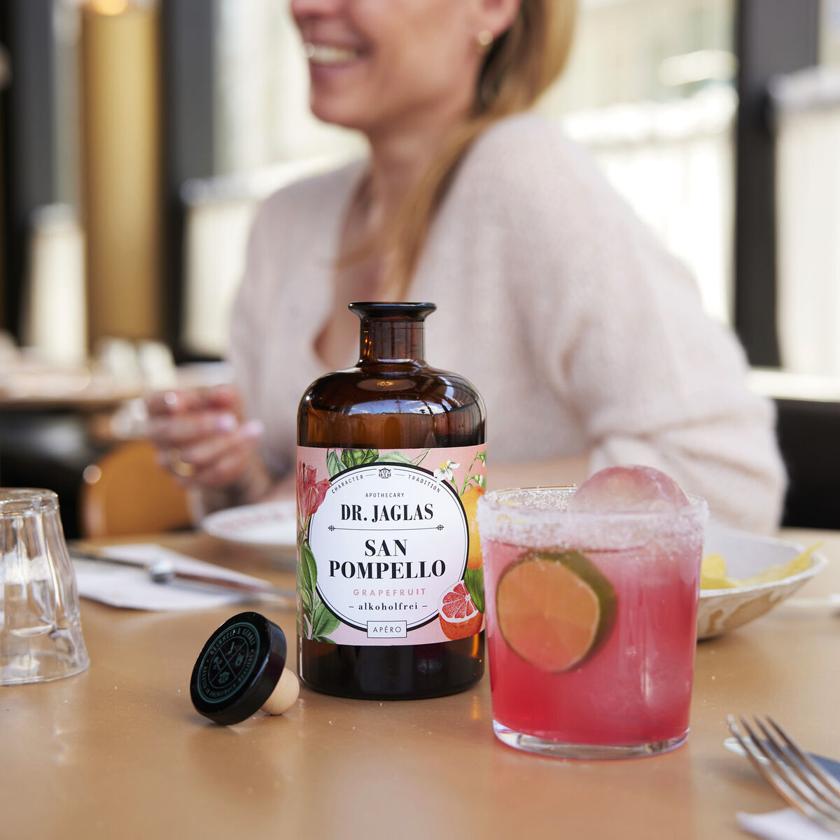 Eine Flasche »San Pomello« alkoholfreier Aperitif steht auf einem Tisch neben einem Glas mit pinkem Getränk, dekoriert mit Limette. Personen sitzen im Hintergrund in einem Restaurantambiente.